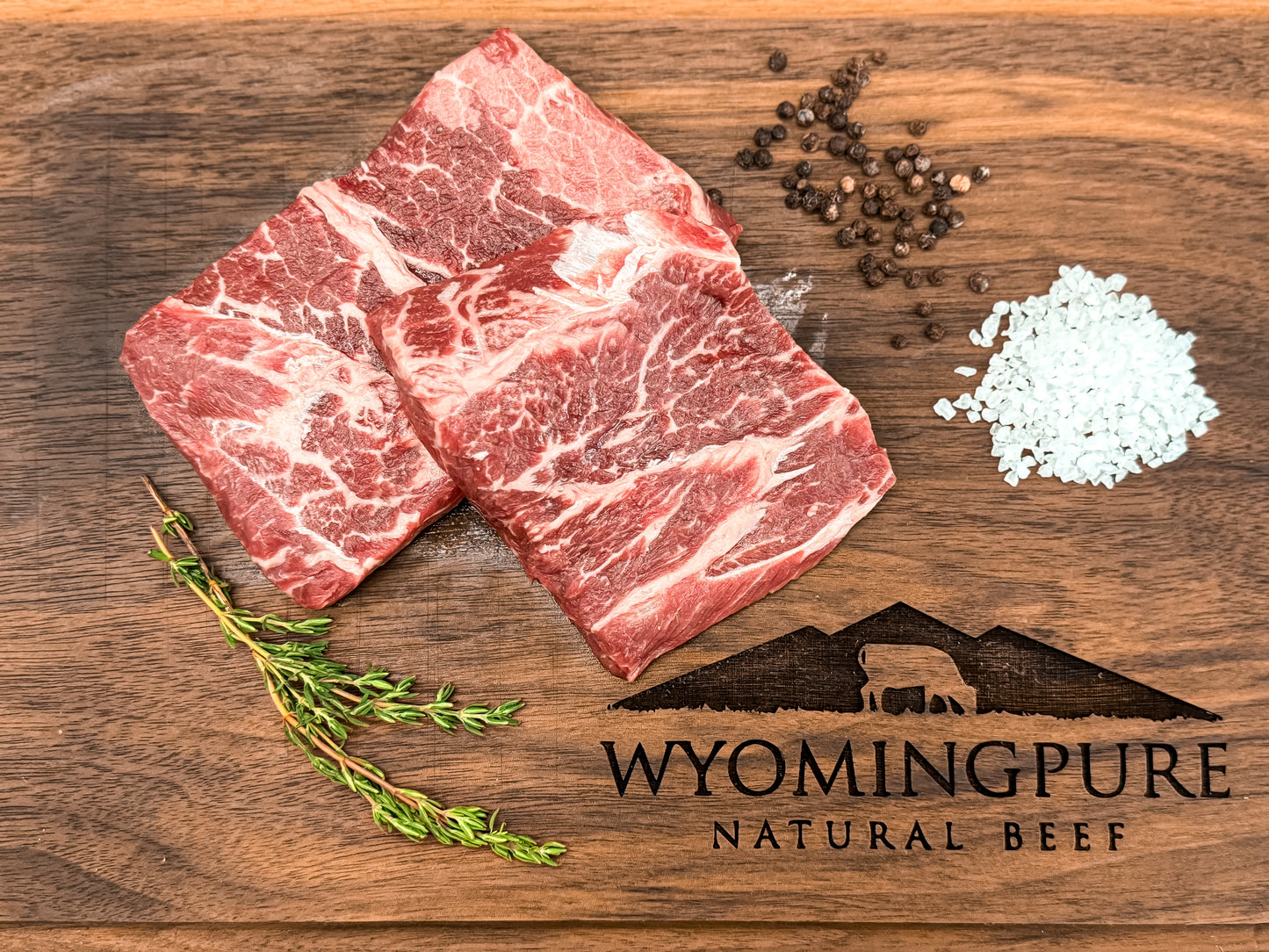 Two raw flat iron steaks on a wooden cutting board with salt, pepper, and a sprig of thyme, displaying the logo of Wyoming Pure Natural Beef.