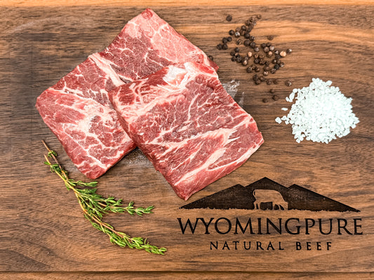 Two raw flat iron steaks on a wooden cutting board with salt, pepper, and a sprig of thyme, displaying the logo of Wyoming Pure Natural Beef.