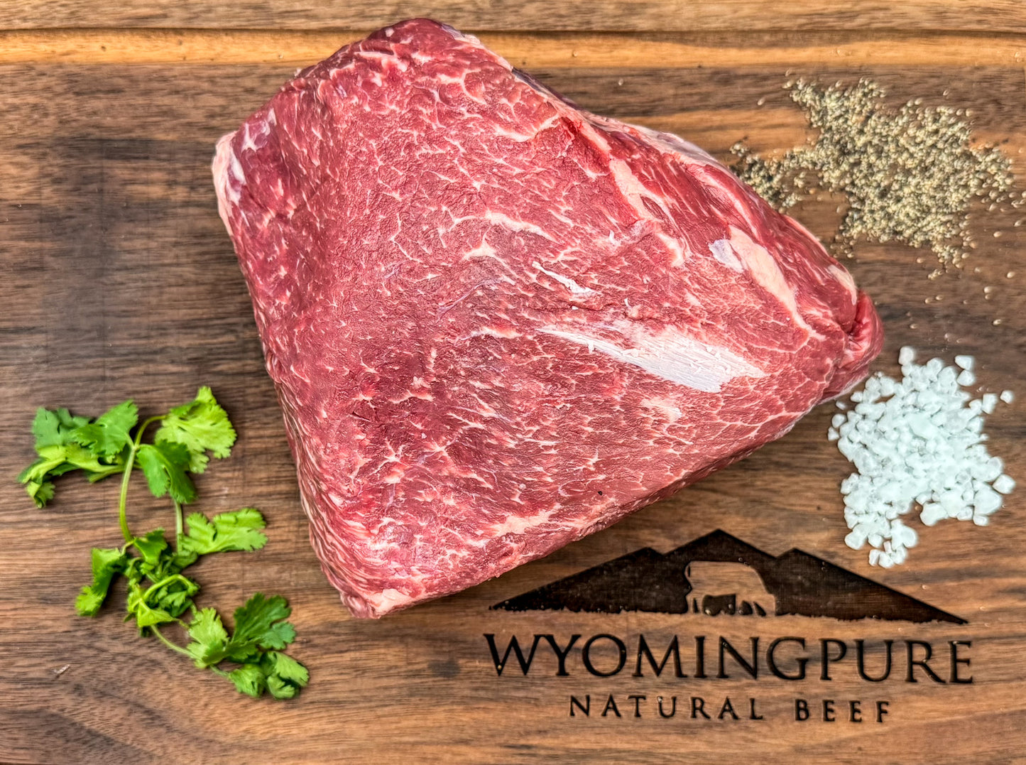 Raw arm roast beef on a wooden board with herbs and spices, branded 'Wyoming Pure Natural Beef'.