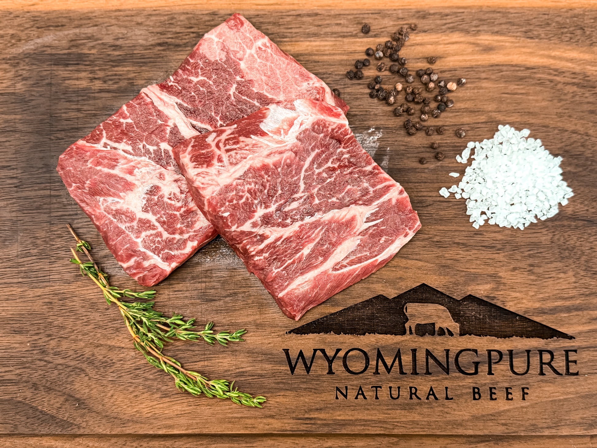 Two raw flat iron steaks on a wooden cutting board with salt, pepper, and a sprig of thyme, displaying the logo of Wyoming Pure Natural Beef.